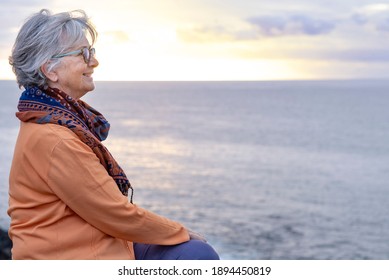 Relaxed Senior Grey-haired Woman Enjoying The Freedom And The Horizon Over Water. Sitting On The Cliff In Front To The Sea In A Winter Cloudy Day