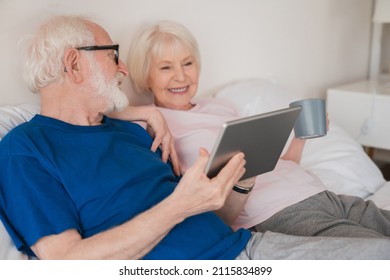 Relaxed senior old elderly couple husband and wife family grandparents resting on the bed together watching films movies on digital tablet, enjoying hot beverage at home. Social distance - Powered by Shutterstock