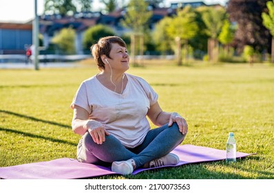 Relaxed senior plus size woman with earphones sitting on yoga mat on green grass outdoors resting after exercises at warm sunny summer day - Powered by Shutterstock