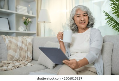 Relaxed senior woman with curly grey hair drinking hot tea and using digital tablet on couch at home. - Powered by Shutterstock