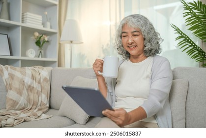 Relaxed senior woman with curly grey hair drinking hot tea and using digital tablet on couch at home. - Powered by Shutterstock