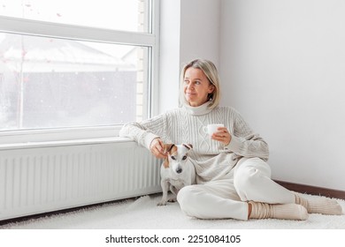 Relaxed, serene adult woman near radiator drinking cup of coffee in the living room. Middle-aged lady sitting with dog on floor at home 