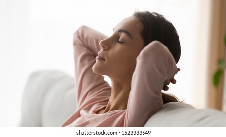Relaxed serene young woman calm face resting on couch breathing fresh air holding hands behind head, healthy girl leaning on sofa napping feel tranquility at home on no stress free weekend, side view