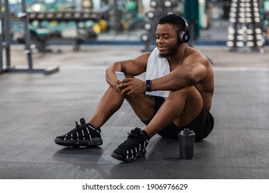 Relaxed shirtless african american sportsman with wireless headset and towel sitting on floor and listening to music on mobile phone, using new musical application for workout, gym interior