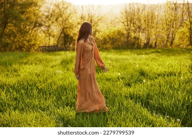 a relaxed slender woman enjoys the sunset standing in a green field with tall grass in an orange dress posing looking away - Shutterstock ID 2279799539