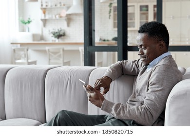 Relaxed smiling african american man holding digital tablet computer using apps sitting on couch at home. Black guy remote learning, social distance working, ordering buying online or reading e book.