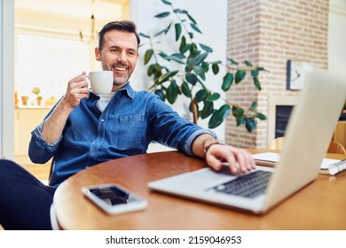 Relaxed smiling man sitting at dinning table at home dinking coffee using laptop                               