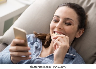 Relaxed Smiling Woman On The Couch At Home, She Is Using A Smartphone And Eating A Snack