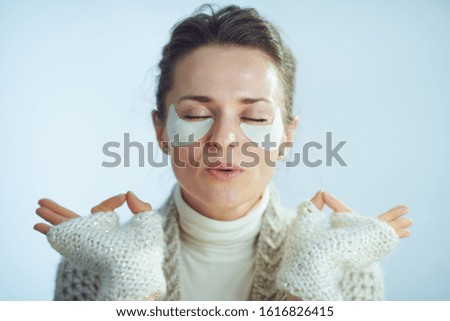 relaxed stylish woman in roll neck sweater and cardigan with cosmetic eye patches meditating against winter light blue background.
