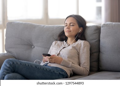 Relaxed teen girl wearing earphones listening to mobile music playing via smartphone app, calm happy young woman in headphones chilling on couch enjoying songs podcast holding cell phone at home