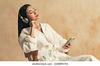 Relaxed teen girl wearing earphones enjoying music or podcast playing via smartphone app, calm happy young korean woman in wireless headphones chilling in morning sitting on chair in home robe - Powered by Shutterstock