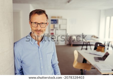 Relaxed thoughtful businessman wearing glasses standing leaning against an office wall scrutinising camera with a quiet smile
