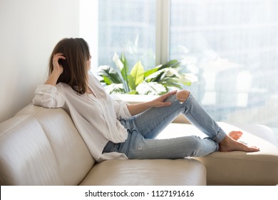 Relaxed Thoughtful Woman Looking Out Of Big Window Enjoying View And Wellbeing Dreaming At Luxury Living Room, Rich Millennial Lady Taking Pleasure Of Stress Free Weekend Resting On Comfortable Sofa