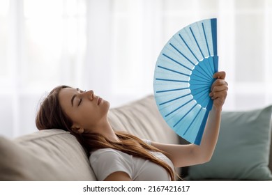 Relaxed tired young european woman with closed eyes suffering from heat on sofa and waving fan at herself in living room interior. Too hot, summer weather and overheating without conditioning at home