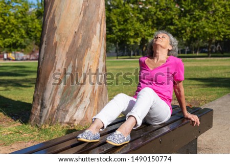 Relaxed tranquil old lady enjoying exercises in city park. Senior grey haired woman in casual sitting and placing feet on bench outdoors, turning face up with closed eyes. Thinking in park concept