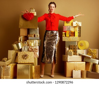 Relaxed Trendy Fashion-monger In Red Blouse And Golden Sparkly Sequin Pencil Skirt With Shopping Bag And Red Heart Doing Yoga Among 2 Piles Of Golden Gifts In Front Of A Plain Wall. Valentine’s Day.