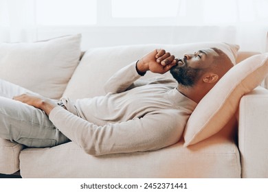 A Relaxed Weekend at Home Smiling African American Man Resting on a Comfortable Sofa in a Modern Living Room He is sitting alone, holding a cup of coffee and thinking The background is filled with a - Powered by Shutterstock