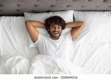 Relaxed well-rested curly bearded millennial indian man laying in comfy bed at weekend, smiling at camera, enjoying new day, wearing pajams, panorama with copy space, top view