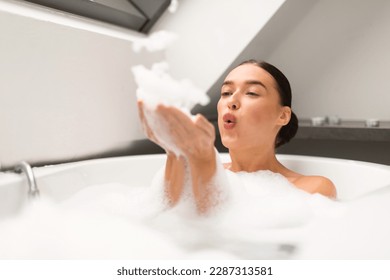 Relaxed Woman Blowing Foam Having Fun Taking Bath Sitting In Bathtub Full Of Hot Water With Soap Bubbles In Bathroom At Home. Lady Enjoying Hygiene Routine Bathing Indoor. Selective Focus