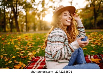 Relaxed woman in a hat enjoys the sunny weather in the park, sitting on a red blanket, drinking a hot drink from a thermos. The concept of rest, relaxation. Lifestyle. - Powered by Shutterstock