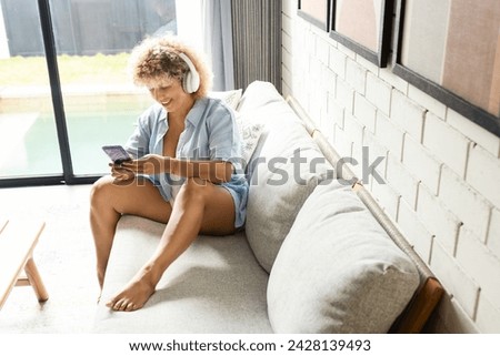 Relaxed Woman with Headphones Using Smartphone on Couch . A cheerful woman enjoys a moment of relaxation on a cozy couch, using her smartphone with white headphones in a brigh