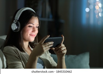 Entspannte Frau mit Kopfhörern, die in der Nacht auf einem Sofa im Wohnzimmer zu Hause auf einem Smartphone Videos guckt