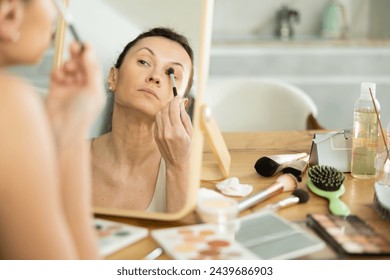 Relaxed woman in her middle age doing make up with brush for eye-shadows looking in the mirror