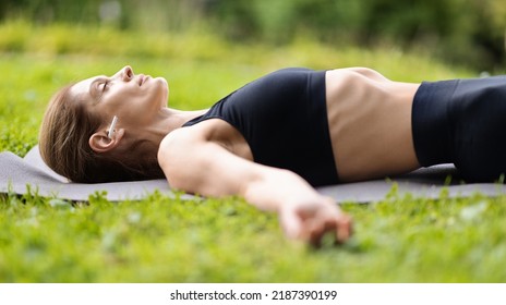 Relaxed Woman Laying On Fitness Mat With Closed Eyes, Meditating And Listening To Music, Using Brand New White Earbuds, Side View, Closeup Shot. Stress Relief, Meditation Outdoors Concept