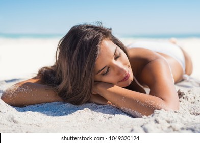 Relaxed woman lying down on sand during summer vacation. Beautiful girl lying down under the sun tanning in a tropical beach. Positive and serene young woman sunbathing at seaside with closed eyes. 