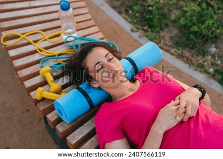 Relaxed woman lying on a bench with yoga mat and exercise equipment after workout, enjoying rest