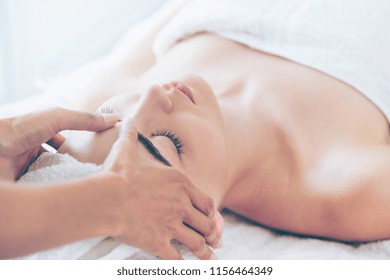 Relaxed Woman Lying On Spa Bed For Facial And Head Massage Spa Treatment By Massage Therapist In A Luxury Spa Resort. Wellness, Stress Relief And Rejuvenation Concept.