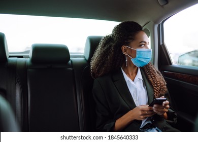 Relaxed woman passenger  in protective medical mask in the taxi car on a backseat with phone. Work on the way to the office. Covid-2019.
