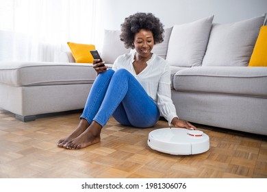 Relaxed woman with phone and robotic vacuum cleaner. Woman using automatic vacuum cleaner to clean the floor, controlling smart machine housework robot with smart phone