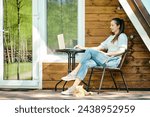Relaxed woman sitting with laptop on terrace of log cabin in a sunny day