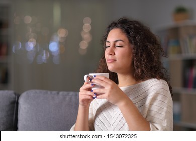 Relaxed woman smelling coffee cup sitting on a couch in the night at home