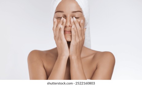 A relaxed woman with a towel wrapped around her head gently massages her face, enjoying a soothing skincare routine. Ideal for promoting beauty, self-care, wellness, and facial care products that