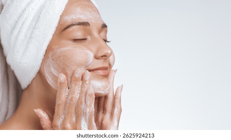 Una mujer relajada con una toalla envuelta alrededor de su cabeza limpia suavemente su cara con jabón espumoso, saboreando su rutina de cuidado de la piel. Perfecto para ilustrar temas de belleza, cuidado personal y piel sana.