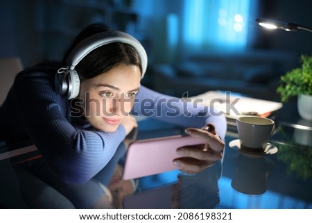 Relaxed woman wearing headphones watching media on smart phone in the night at home