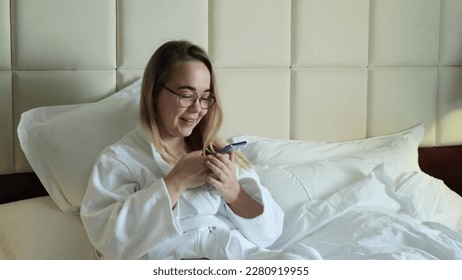 A relaxed woman in a white coat sits on a mole in a hotel room and records a voice message on her smartphone. Online communication. Application or messenger. - Shutterstock ID 2280919955