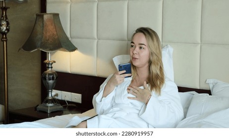 A relaxed woman in a white coat sits on a mole in a hotel room and records a voice message on her smartphone. Online communication. Application or messenger. - Shutterstock ID 2280919957