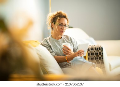 Relaxed young adult caucasian woman read a book on modern device reader comfortably sitting on the couch at home. Portrait of pretty female people wearing eyeglasses and using connection - Powered by Shutterstock