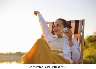 Relaxed Young Adult Happy Satisfied Caucasian Female Camper Sitting On A Camping Chair And Having A Morning Coffee Near The River. Camping And Outdoor Activity.