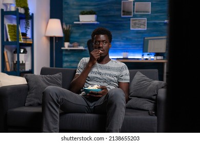 Relaxed Young Adult Resting On Couch Watching Football Game On Television Spending Evening Alone At Home In Living Room. Guy Holding Popcord Bowl Looking At Comedy Movie On Tv Enjoying Free Time