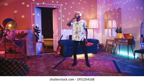 Relaxed Young African American Male In Living Room Listening To Music In Headphones And Dancing, Moving Body Rhythmically In Neon Disco Light In Vintage Apartment. Retro Style, Leisure Concept