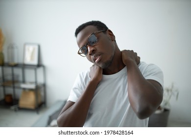 Relaxed young Afro American male wearing spectacles and t-shirt keeping eyes closed, massaging neck after awakening, relaxing muscles. Black guy doing warm physical exercises, rotating head - Powered by Shutterstock