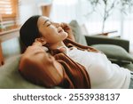 Relaxed young Asian woman enjoying rest on comfortable sofa, calm attractive girl relaxing on couch, breathing fresh air with eyes closed, meditating at home