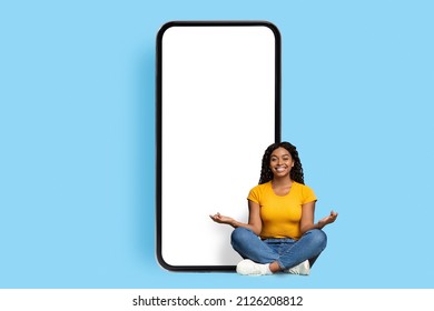 Relaxed young black woman sitting on floor in lotus pose, meditating by huge mobile phone with white blank screen, using sports mobile app, blue studio background, mockup, copy space
