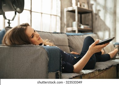 Relaxed Young Brunette Woman Lying On Sofa And Watching Tv In Loft Apartment. Urban Chic Loft Decoration Details And Window. Modern Lifestyle Concept.