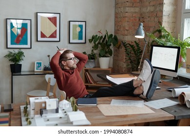 Relaxed young Caucasian bearded architect in eyeglasses sitting with feet on desk and looking at monitor with sketch in office