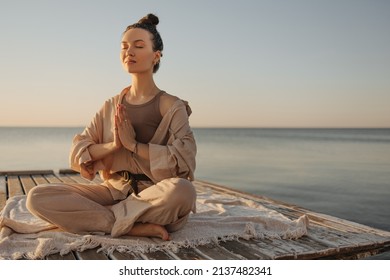 Relaxed young caucasian woman sitting on seashore practices yoga without stress. Model with dark topknot on her head with eyes closed. Cozy beach atmosphere, summer concept. - Powered by Shutterstock